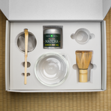 Hario and Mizuba Matcha tea gift set with a clear glass bowl, bamboo chasen whisk, bamboo chashaku scoop, and metal matcha sieve in a box on a tatami mat surface.