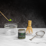 Matcha tea gift set with a tin of Mizuba Okumidori single cultivar matcha. A bamboo chashaku spoon with matcha hovers over a glass chawan tea bowl that's next to a bamboo chasen whisk and metal sieve on a marble surface.