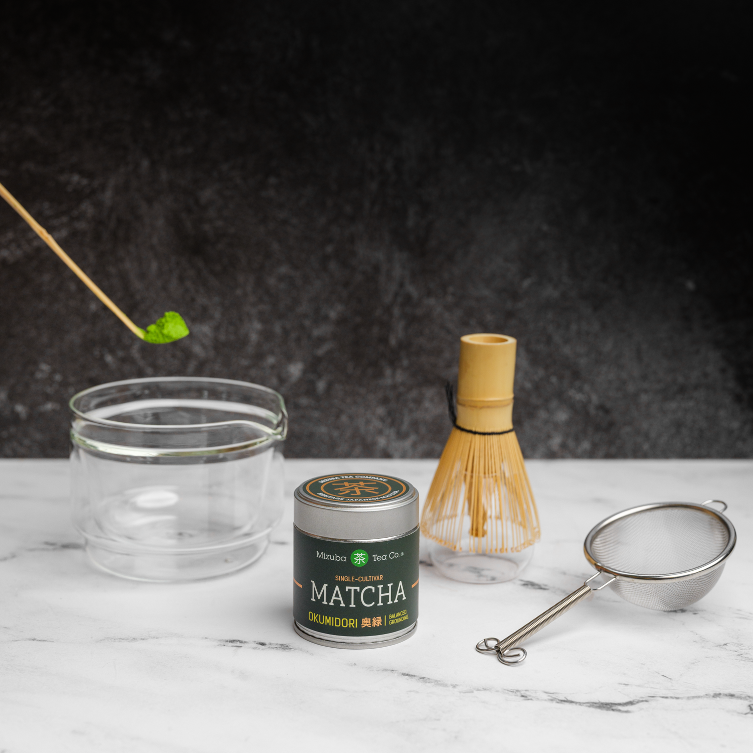 Matcha tea gift set with a tin of Mizuba Okumidori single cultivar matcha. A bamboo chashaku spoon with matcha hovers over a glass chawan tea bowl that's next to a bamboo chasen whisk and metal sieve on a marble surface.