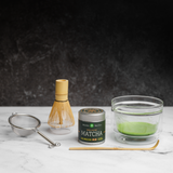 Matcha green tea set complete with traditional tools. A glass chawan bowl with frothy matcha green tea inside sits next to a whisk, a bamboo chashaku scoop, a metal sieve, and a Mizuba Tea Okumidori matcha tin on a dark background.