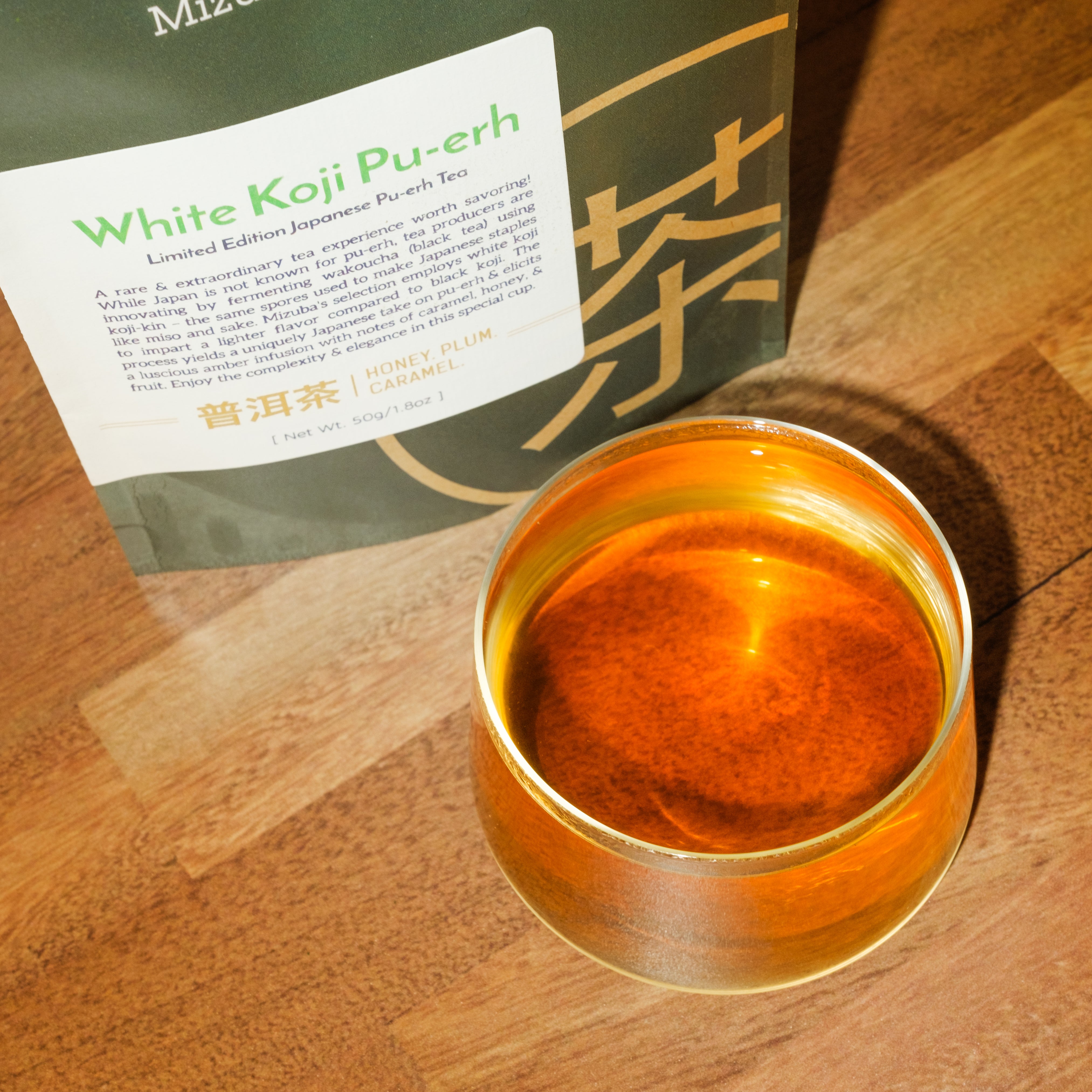 An overhead view showcases amber pu-erh tea in a glass tea cup. The tea's packaging is in the background – Mizuba Tea Co.