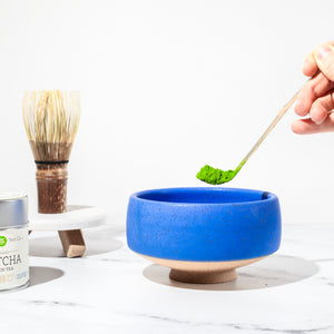 A hand places a scoop of pure Japanese matcha into a cerulean matcha chawan (tea bowl). In the background is a dark bamboo whisk and matcha tin.