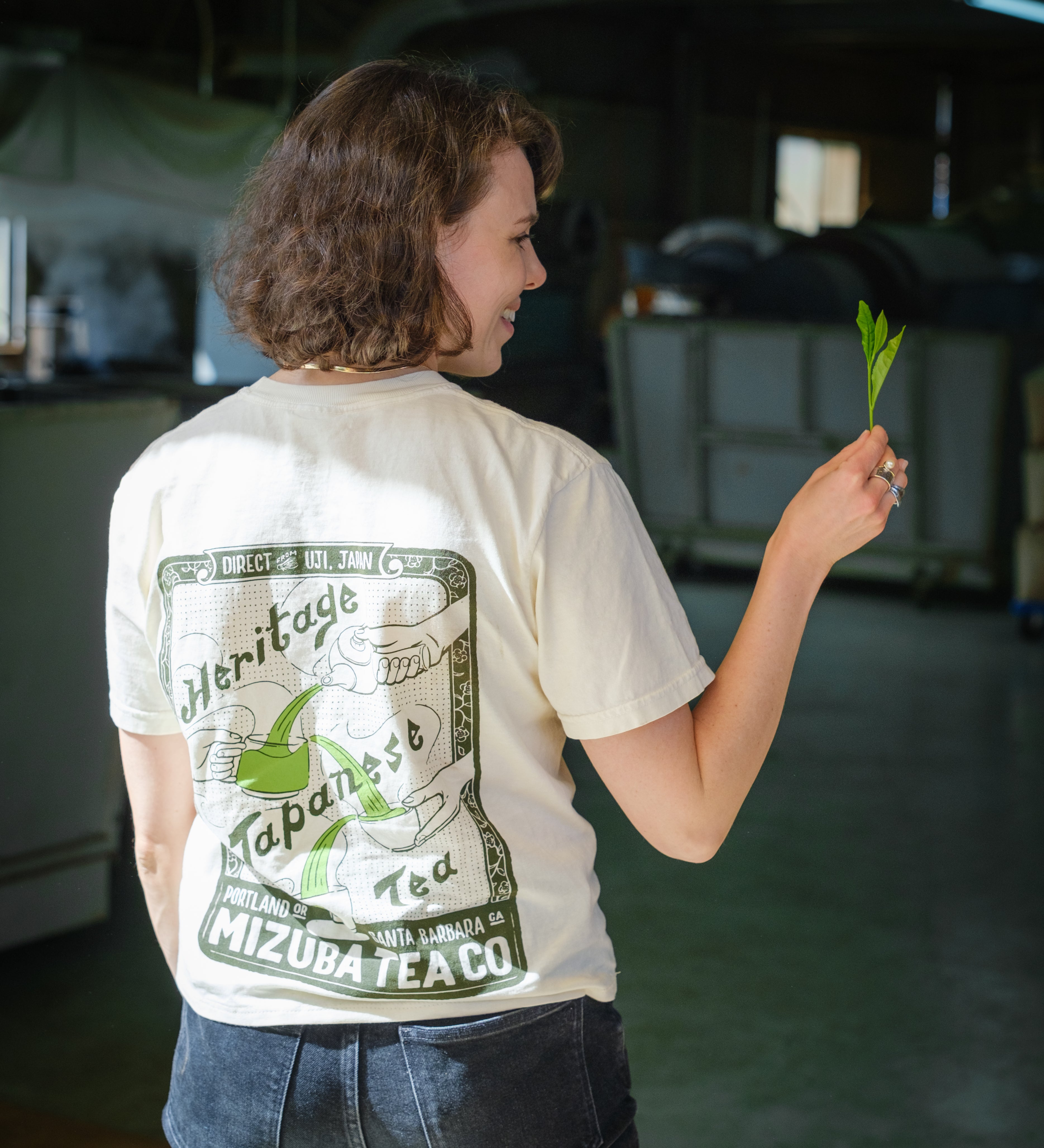 Woman wearing a custom illustrated shirt with Japanese tea design holds a fresh tea leaf from the fields of Japan