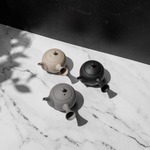 Three ceramic Japanese kyusu teapots in beige, black, and gray on a marble surface with shadows.