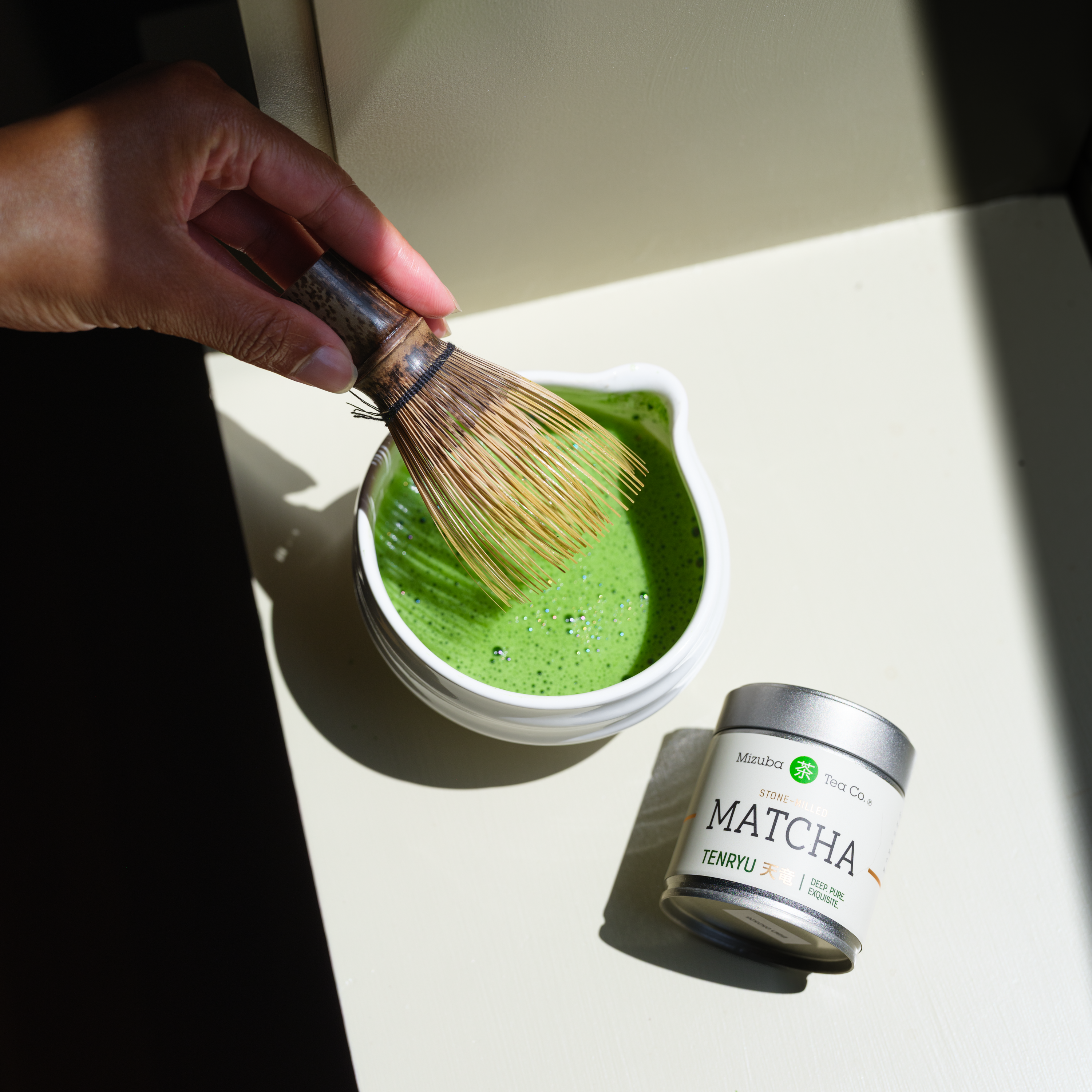Matcha green tea powder being whisked in a chawan tea bowl with a chasen matcha whisk, next to a can of Mizuba matcha powder.
