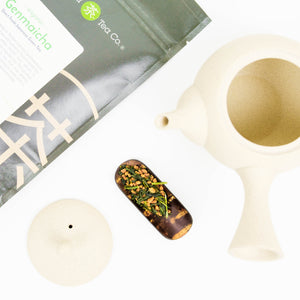 Tea items lay on a white surface. A bag of genmaicha tea is featured next to a wooden teascoop and white, Japanese tea pot