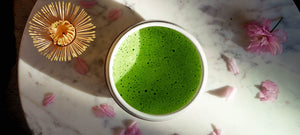 A frothy bowl of matcha sits on a marble table top covered in cherry blossom petals. A chasen bamboo tea whisk is to the left of the bowl.