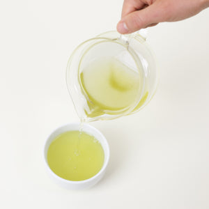Hand pours Japanese sencha green tea into a white cup on a white table top