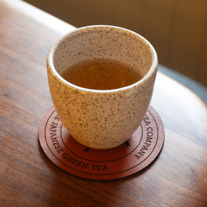A white teacup is placed on top of a leather coaster that says Japanese Green Tea on a wooden table.