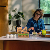 A woman in a blue shirt stands in a kitchen behind a counter, whisking matcha. On the counter are several small silver jars labeled MATCHA and a decorative illustrated box, with a bright green cup beside her. Product featured is the Mizuba Tea Ceremonial Matcha Tasting Trio Set.