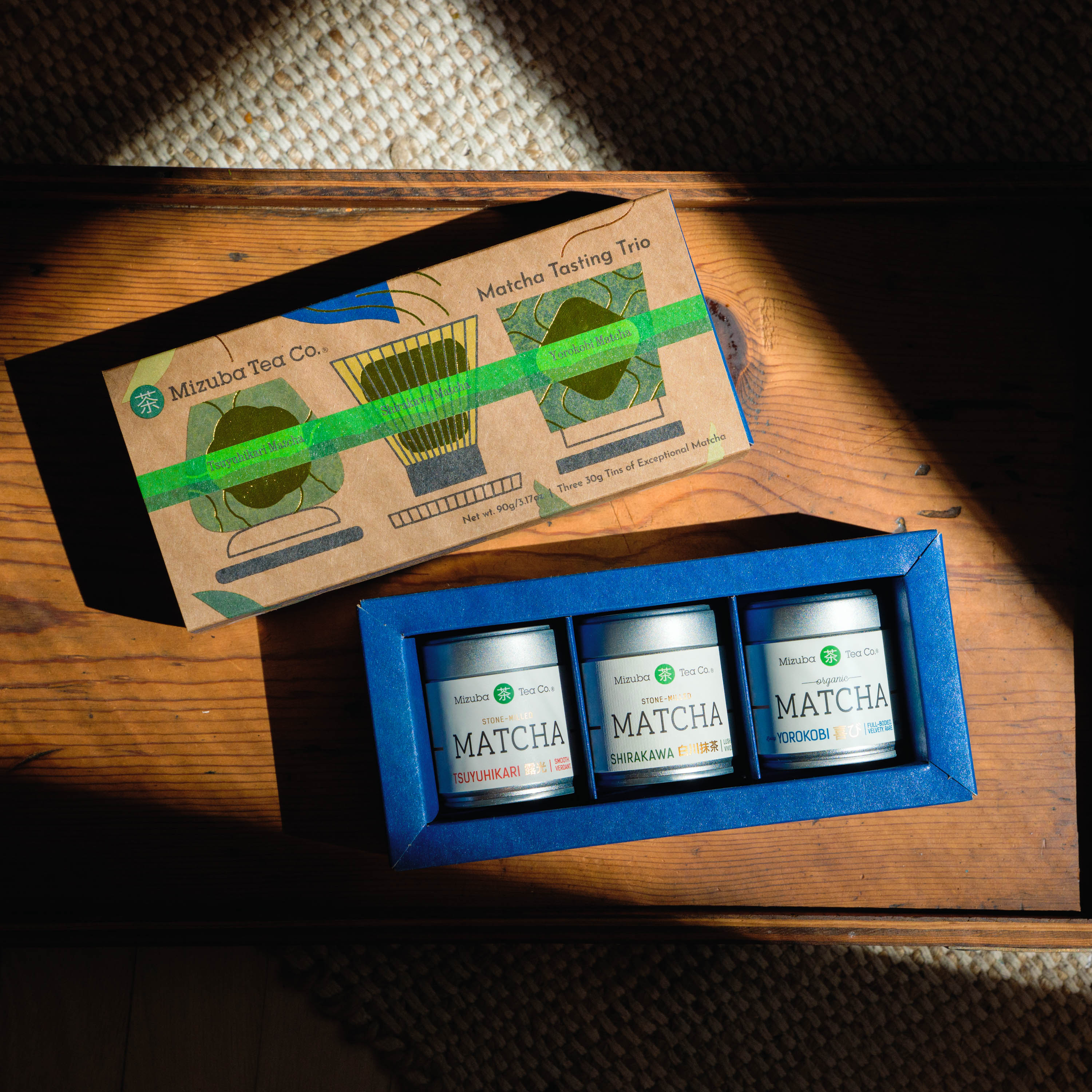 A ceremonial matcha tasting set from Mizuba Tea Co. displayed on a wooden surface in natural sunlight. The three tins read, "Tsuyuhikari, Shirakawa, and Yorokobi Matcha."