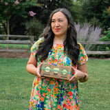A woman with long dark hair stands outdoors on a grassy lawn holding a Mizuba Ceremonial Matcha Tasting Set. She is wearing a colorful floral dress, with trees and a wooden fence in the background.