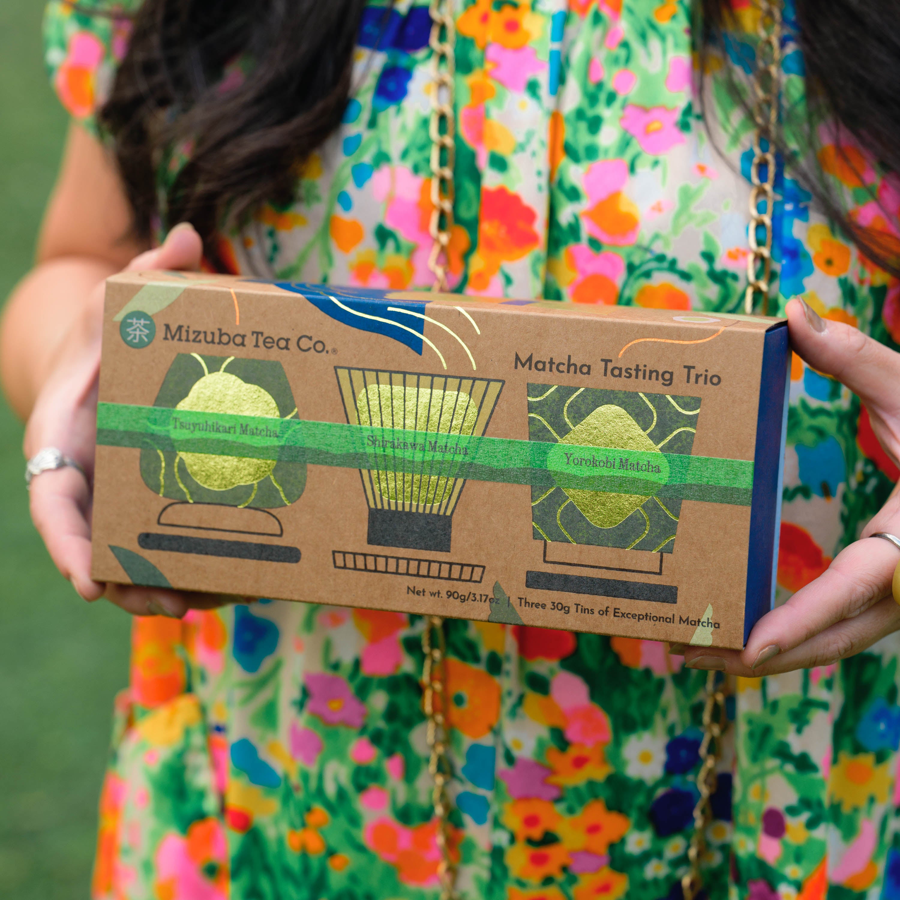 A woman in a colorful floral dress holds a box labeled Mizuba Tea Co. Matcha Tasting Trio. 