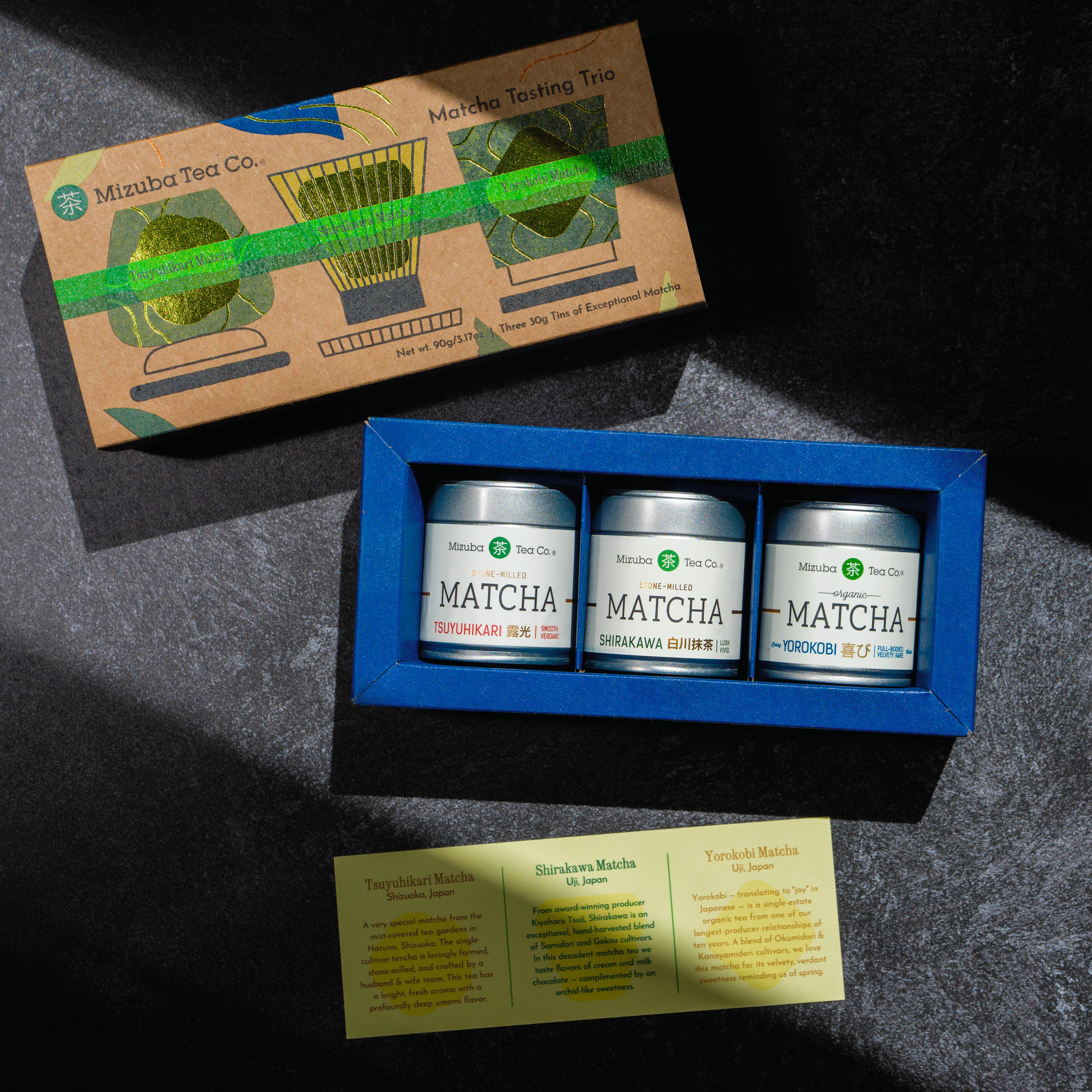 A Mizuba Tea Co. Matcha Tea Tasting Trio, featuring Tsuyuhikari, Shirakawa, and Yorokobi tins, displayed on a dark textured surface. Includes tasting notes.