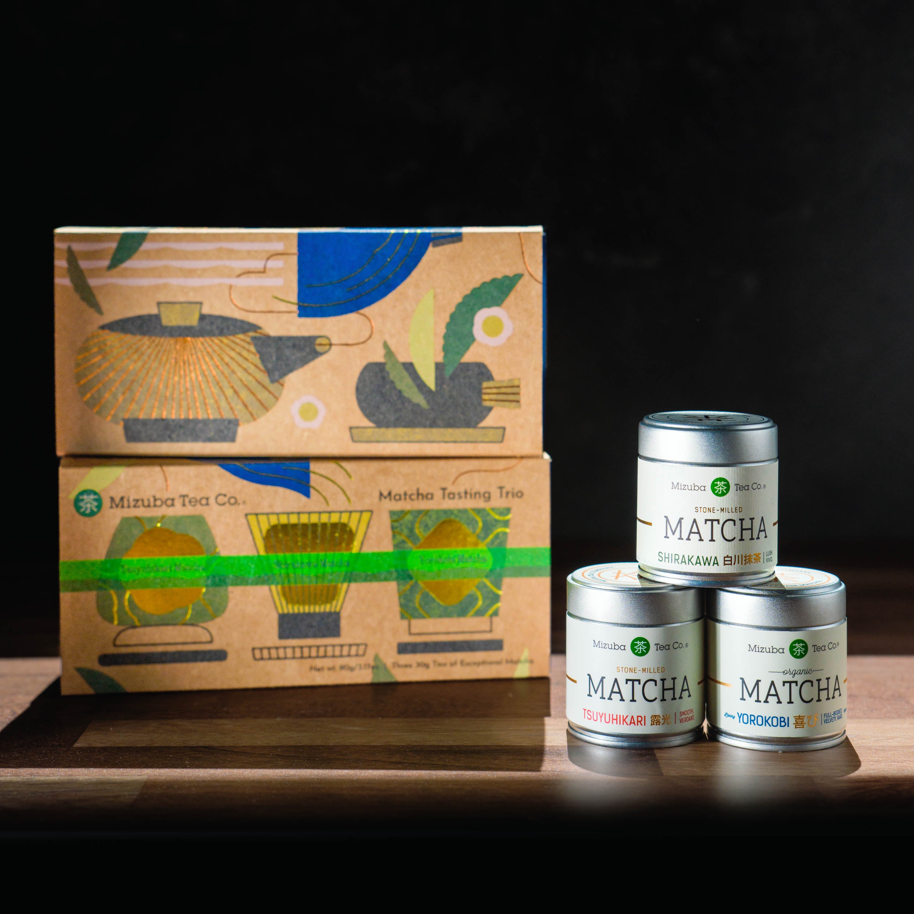 A boxed set of Mizuba Tea Co. ceremonial matcha tea alongside three small silver matcha tins on a wooden surface against a dark background.