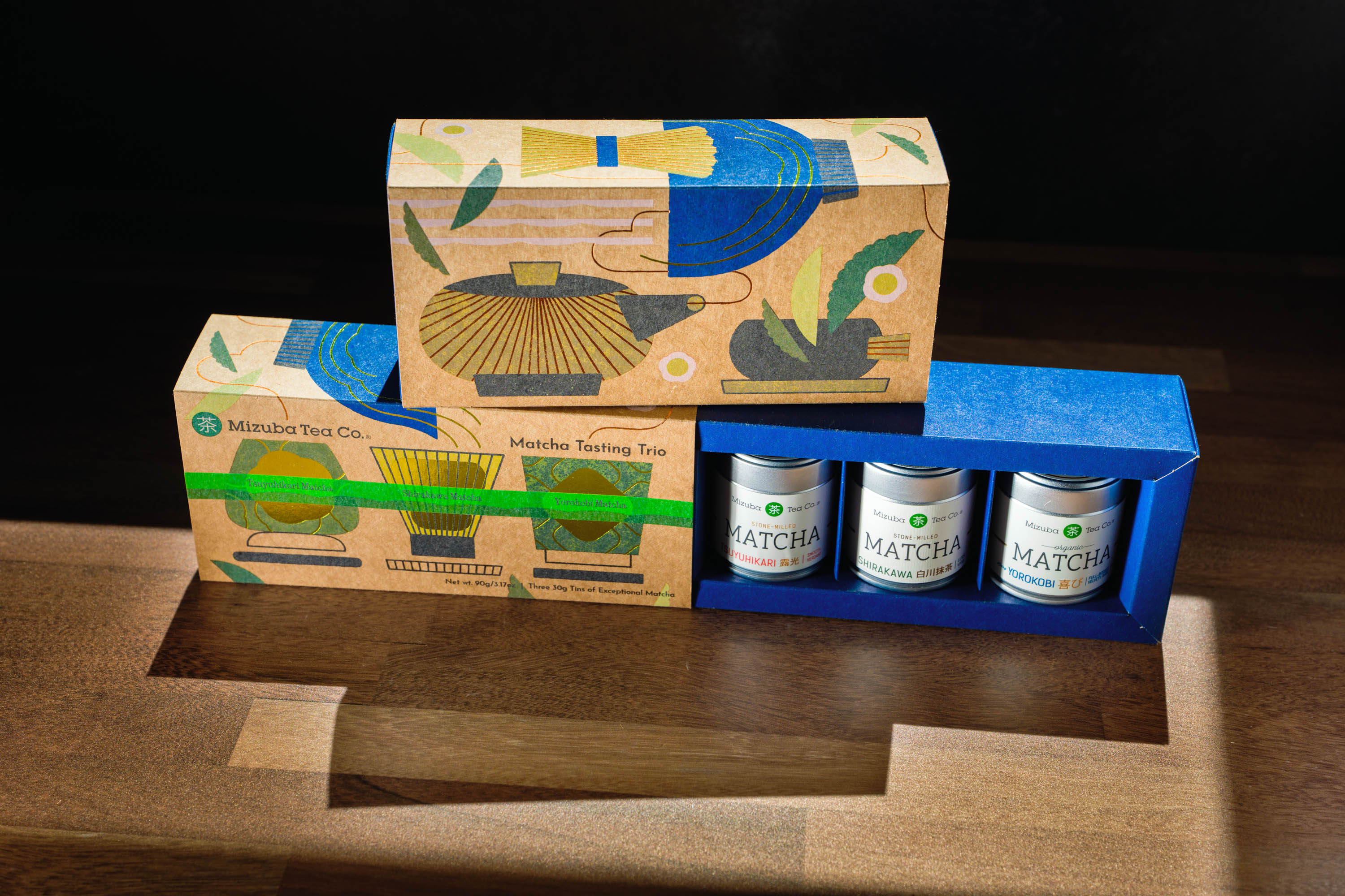 A boxed matcha tea gift set by Mizuba Tea Co is displayed on a wooden surface against a dark background. The set includes an artistic, illustrated box holding three silver matcha tins labeled Tsyuhikari, Shirakawa, and Yorokobi MATCHA.