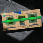 A kraft paper box labeled 'Mizuba Tea Co. Matcha Tasting Trio' photographed on a dark textured surface with dramatic shadow lighting. Three ceremonial matcha teas featured are Tsuyuhikari, Shirakawa, and Yorokobi organic matcha.