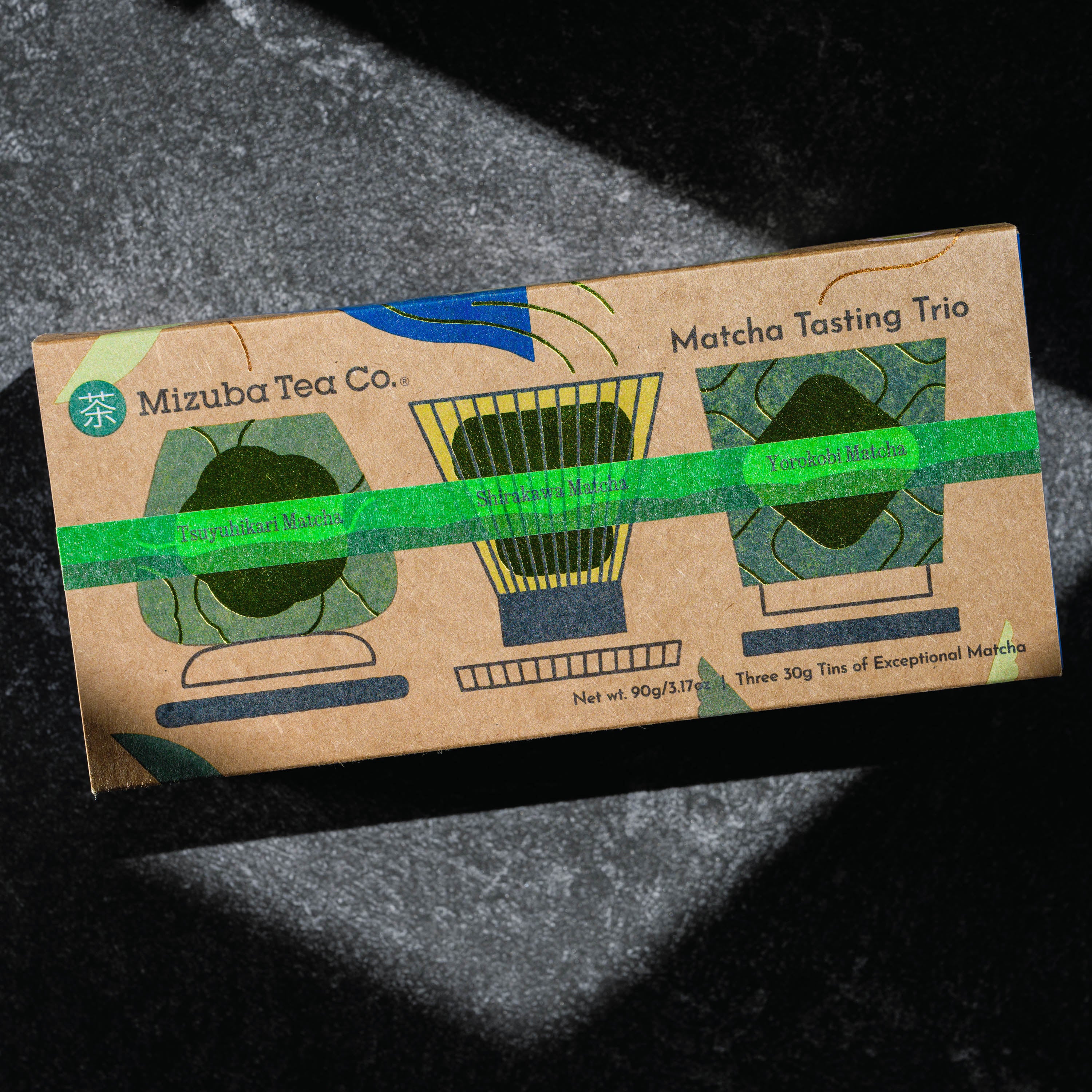 A kraft paper box labeled 'Mizuba Tea Co. Matcha Tasting Trio' photographed on a dark textured surface with dramatic shadow lighting. Three ceremonial matcha teas featured are Tsuyuhikari, Shirakawa, and Yorokobi organic matcha.
