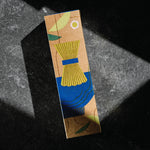 A rectangular kraft paper box with an abstract illustration of a chasen matcha bamboo whisk in yellow, blue, green, and brown tones, photographed on a dark textured surface with dramatic diagonal lighting. Product by Mizuba Tea Co.