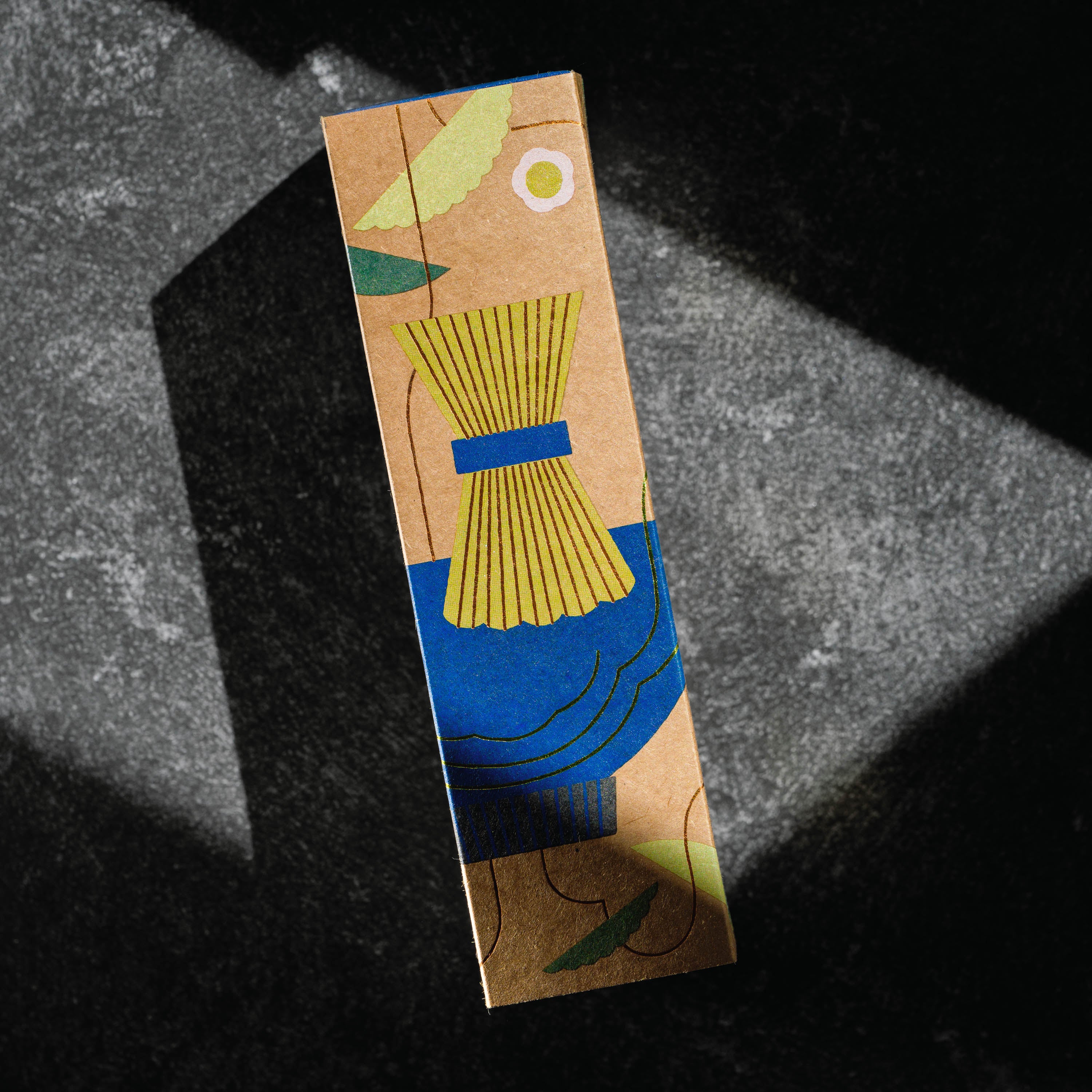 A rectangular kraft paper box with an abstract illustration of a chasen matcha bamboo whisk in yellow, blue, green, and brown tones, photographed on a dark textured surface with dramatic diagonal lighting. Product by Mizuba Tea Co.