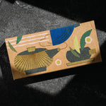Matcha set by Mizuba Tea Co. A rectangular kraft paper box with an abstract illustrated design of a teapot and a tea cup in blue, green, gold, and dark gray, photographed on a dark textured surface with dramatic shadows. 