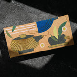 Matcha set by Mizuba Tea Co. A rectangular kraft paper box with an abstract illustrated design of a teapot and a tea cup in blue, green, gold, and dark gray, photographed on a dark textured surface with dramatic shadows. 