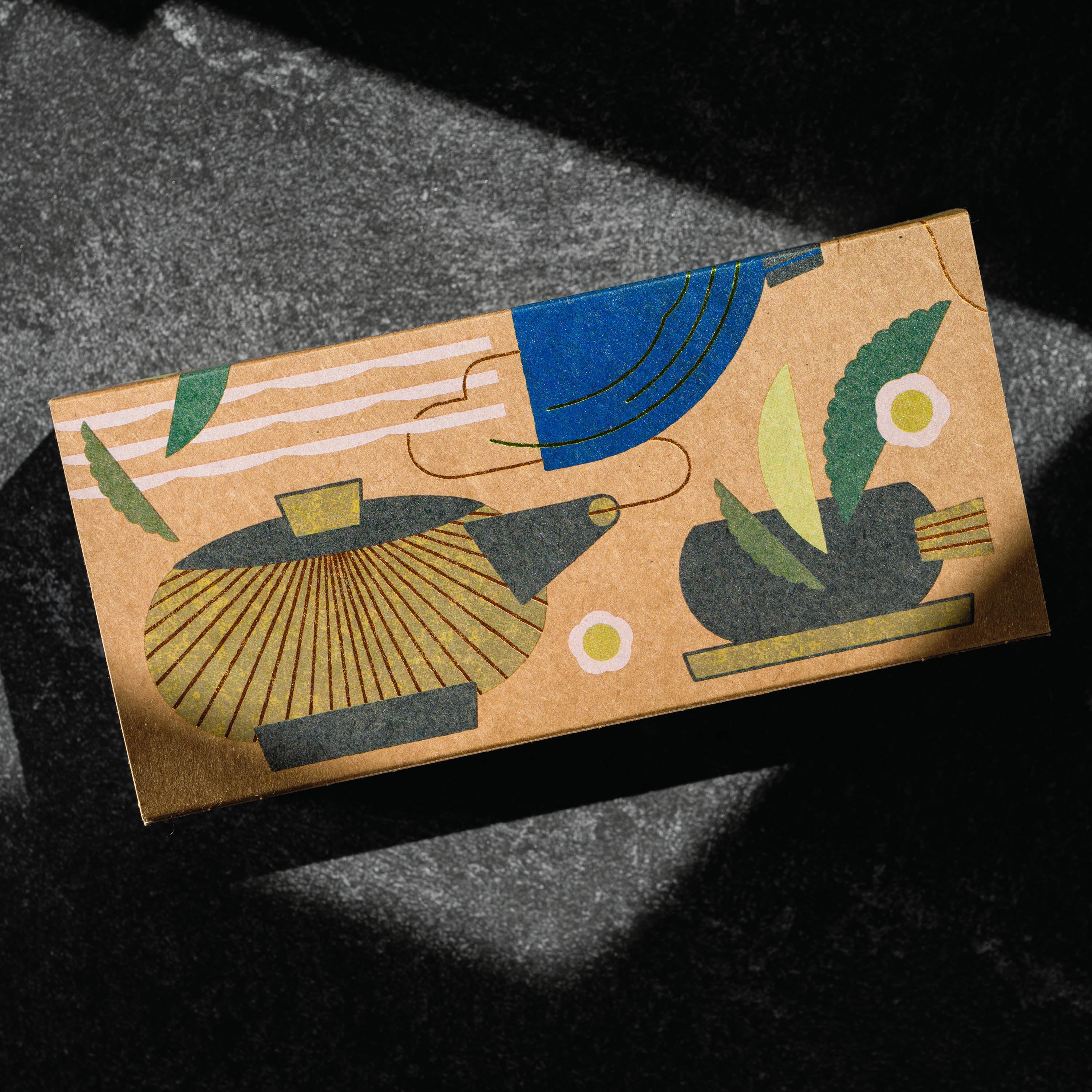 Matcha set by Mizuba Tea Co. A rectangular kraft paper box with an abstract illustrated design of a teapot and a tea cup in blue, green, gold, and dark gray, photographed on a dark textured surface with dramatic shadows. 