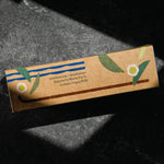 A rectangular kraft paper box for matcha green tea, featuring blue stripes, green leaves, and white flower illustrations, photographed on a dark textured surface with dramatic shadows. By Mizuba Tea Co.