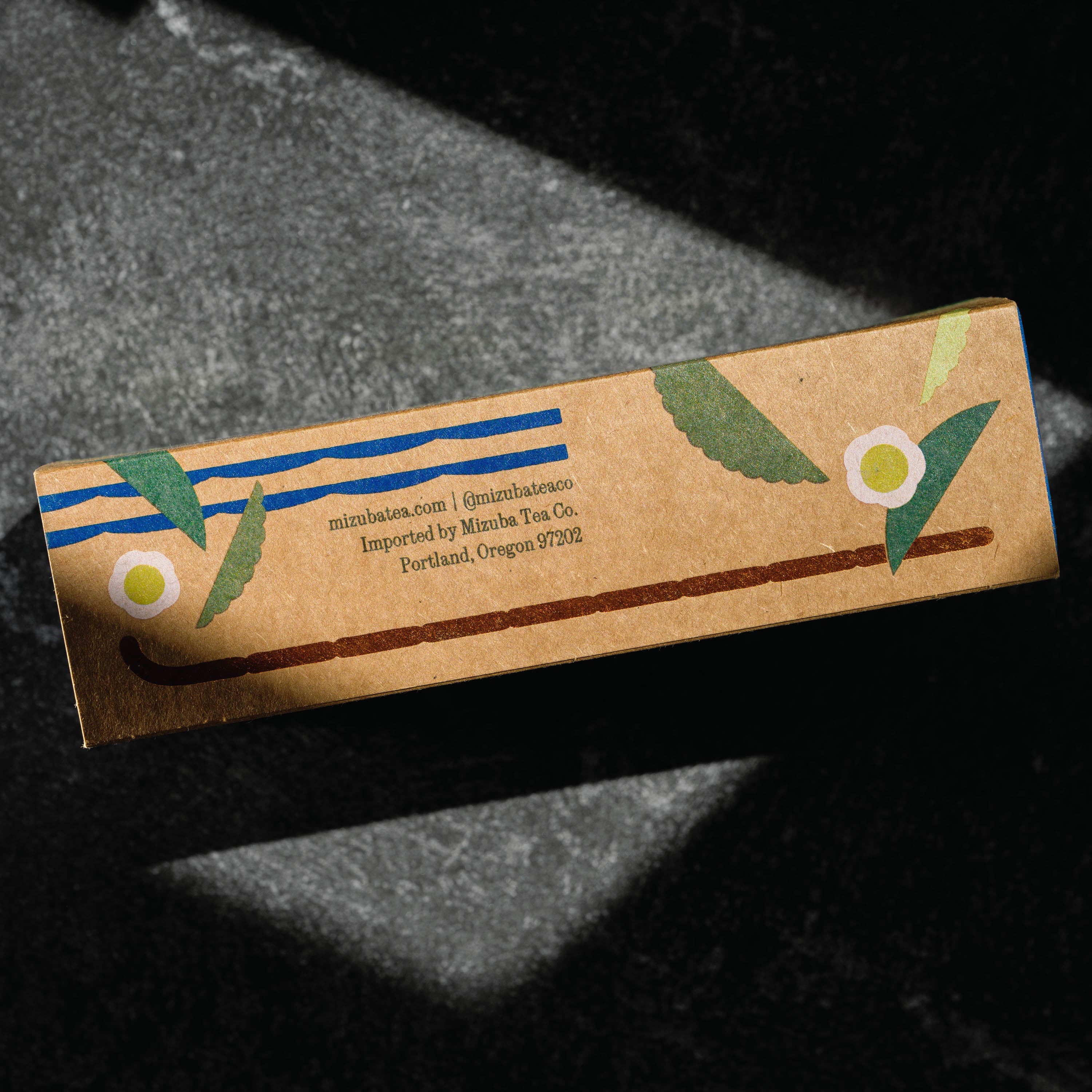 A rectangular kraft paper box for matcha green tea, featuring blue stripes, green leaves, and white flower illustrations, photographed on a dark textured surface with dramatic shadows. By Mizuba Tea Co.