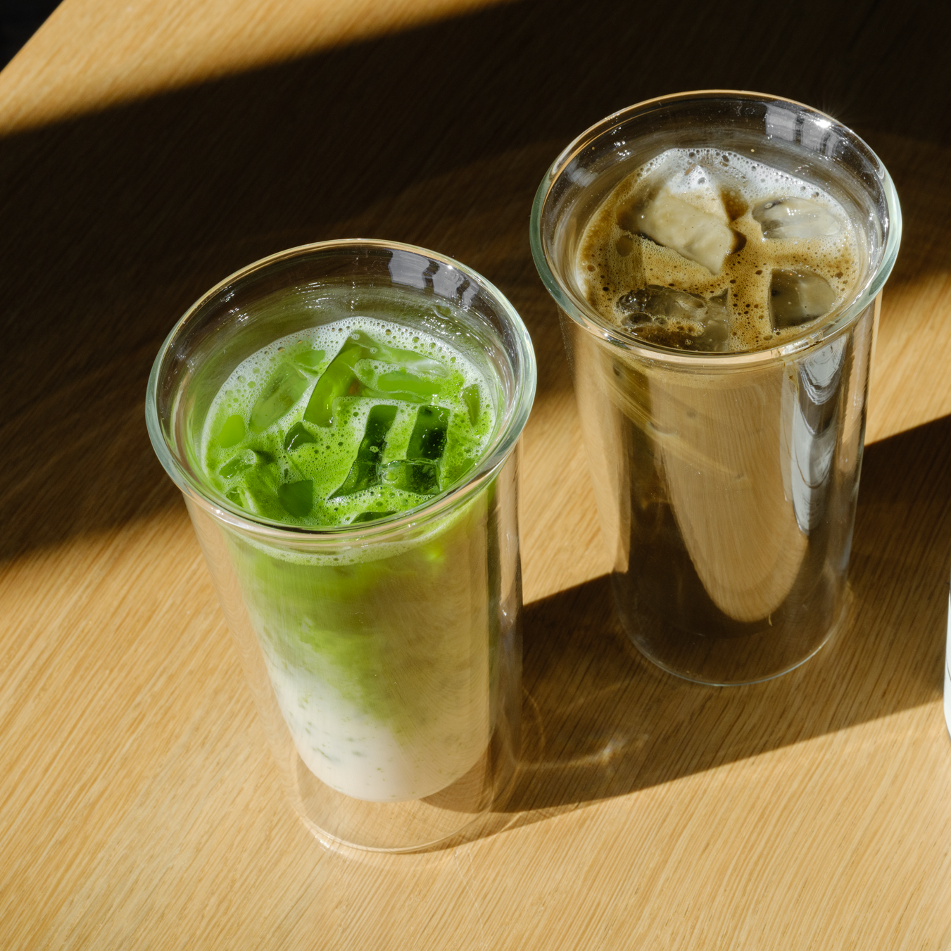 One glass of an iced Mizuba matcha latte on the left stands next to an iced Mizuba hojicha latte on the right. 