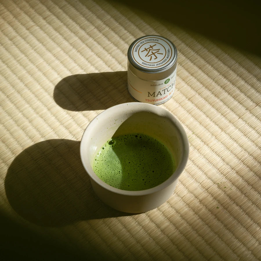 A bowl of frothy matcha green tea sits next to a tin of single cultivar, Tsuyuhikari Japanese matcha on tatami mat. 