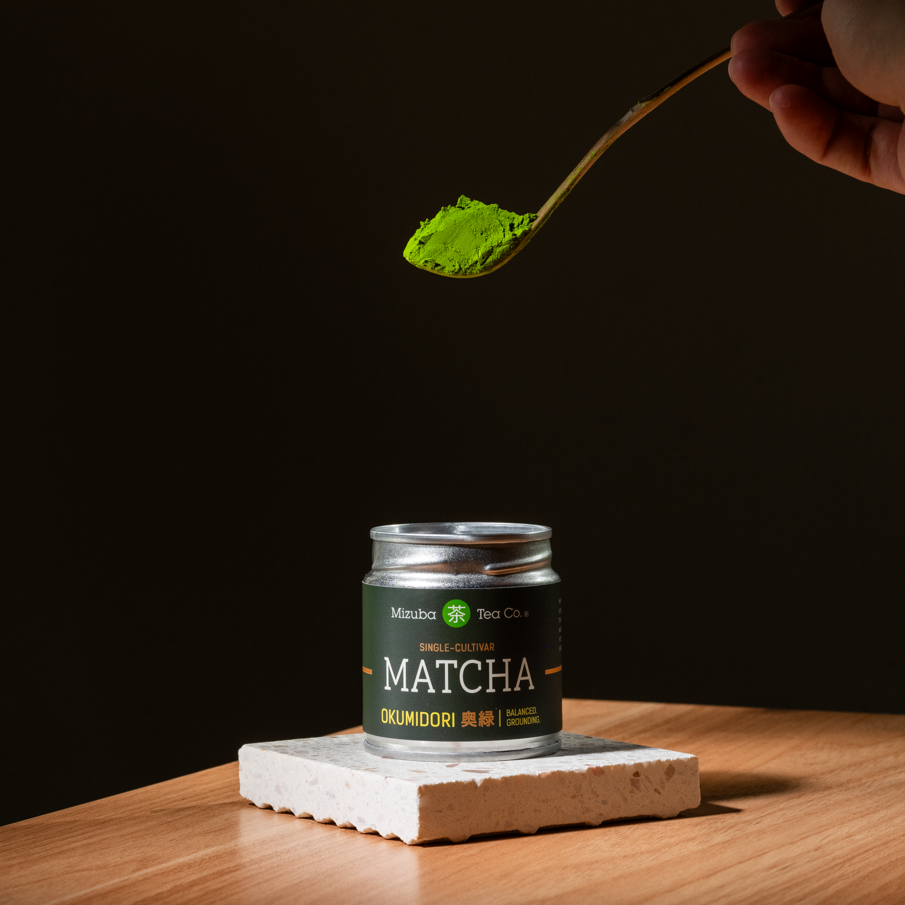 Jar of matcha green tea powder with a spoonful being held above it on a dark background