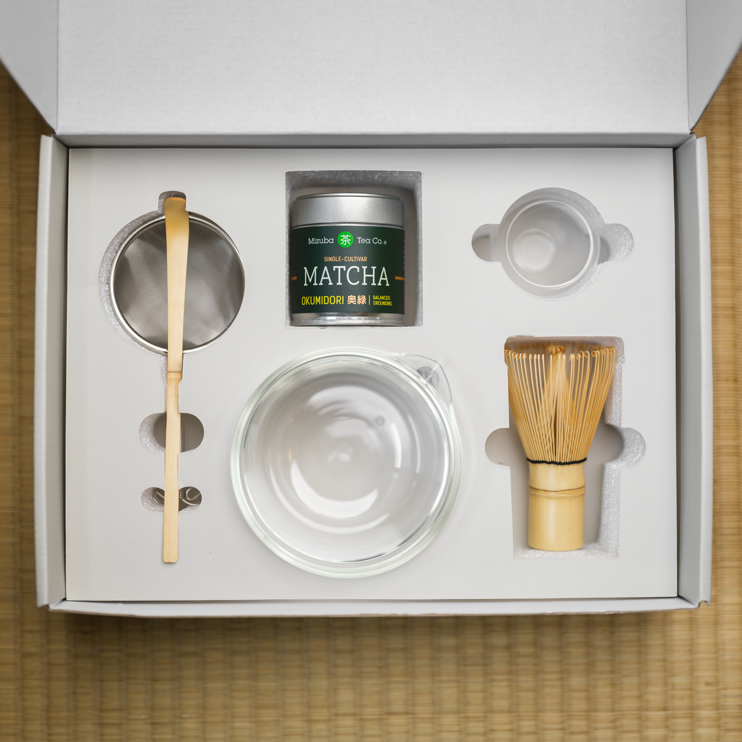 Hario and Mizuba Matcha tea gift set with a clear glass bowl, bamboo chasen whisk, bamboo chashaku scoop, and metal matcha sieve in a box on a tatami mat surface.