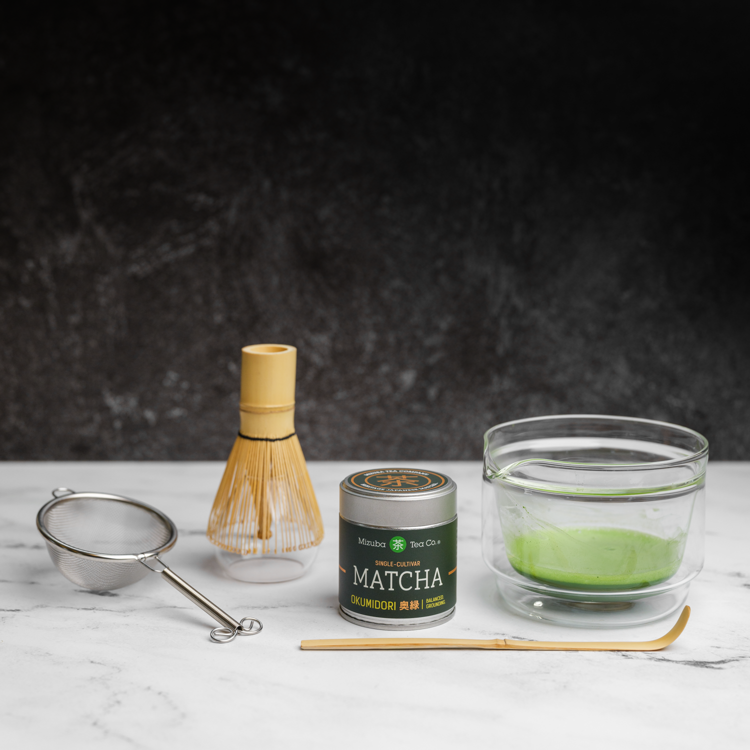 Matcha green tea set complete with traditional tools. A glass chawan bowl with frothy matcha green tea inside sits next to a whisk,  a bamboo chashaku scoop, a metal sieve, and a Mizuba Tea Okumidori matcha tin on a dark background.