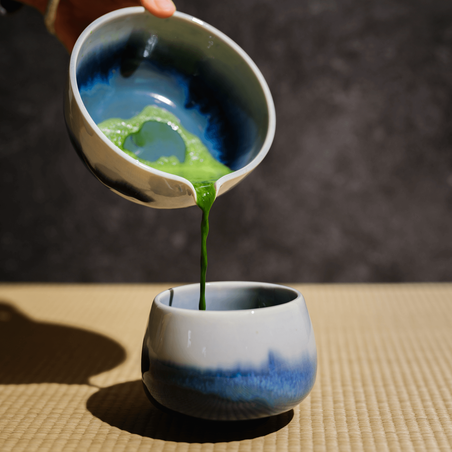 Freshly whisked matcha green tea poured from a ceramic chawan into a blue gradient cup on a tatami mat.