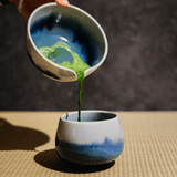Freshly whisked matcha green tea poured from a ceramic chawan into a blue gradient cup on a tatami mat.
