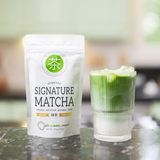 Bag of Mizuba Tea Co. Organic Japanese Signature Matcha tea and a glass of matcha green tea latte on a reflective surface.