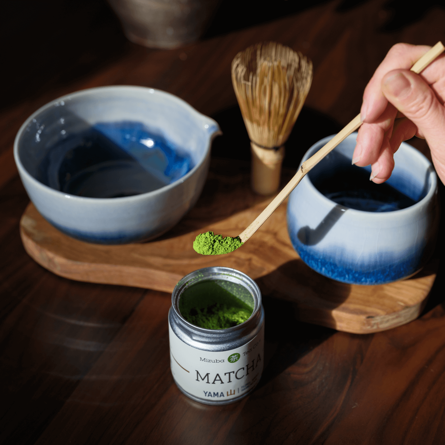Person preparing matcha tea with a container of matcha powder on a wooden table. A blue and white chawan, bamboo chasen, and blue ceramic cup are also on the table.