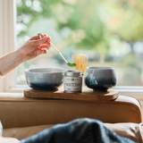 Person making matcha tea with two bowls and a Mizuba Tea Co. Yama Matcha tin on a wooden tray by a window.