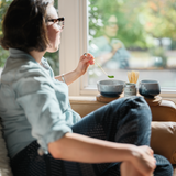 Woman sitting by a window enjoying  preparing a bowl of matcha green tea using a traditional, Japanese bamboo chashaku by Mizuba Tea Company. 