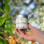 Hand holding a canister of Matcha Yama tea from Matcha Tea Co. with a blurred natural background.