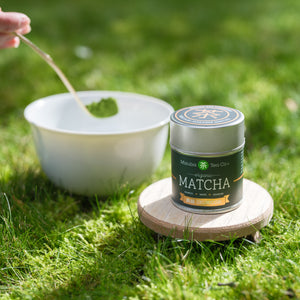 A dark green tin of organic Mizuba Matcha sits in front of a porcelain white tea bowl with a scoop of matcha about to be placed inside.
