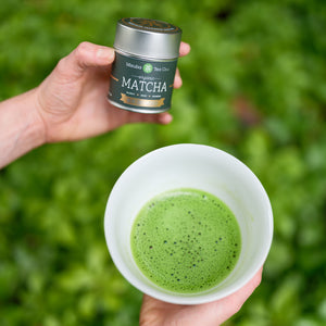 A hand holds a dark green tin of Mizuba Tea Co. Organic Okumidori matcha green tea. The other hand holds a white chawan tea bowl filled with frothy green whisked matcha. 