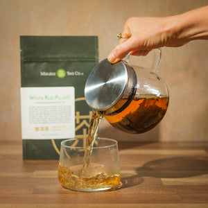 A hand pours a glass teapot of Japanese pu-erh fermented with koji into a glass tea cup on a wooden surface. In the background is a Mizuba Tea Co. branded bag.