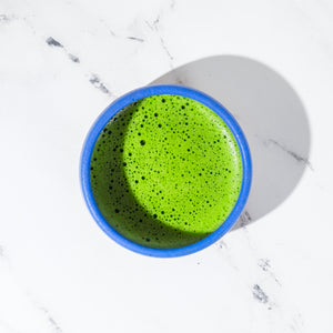 A blue bowl of frothy matcha sits on top of a marble surface