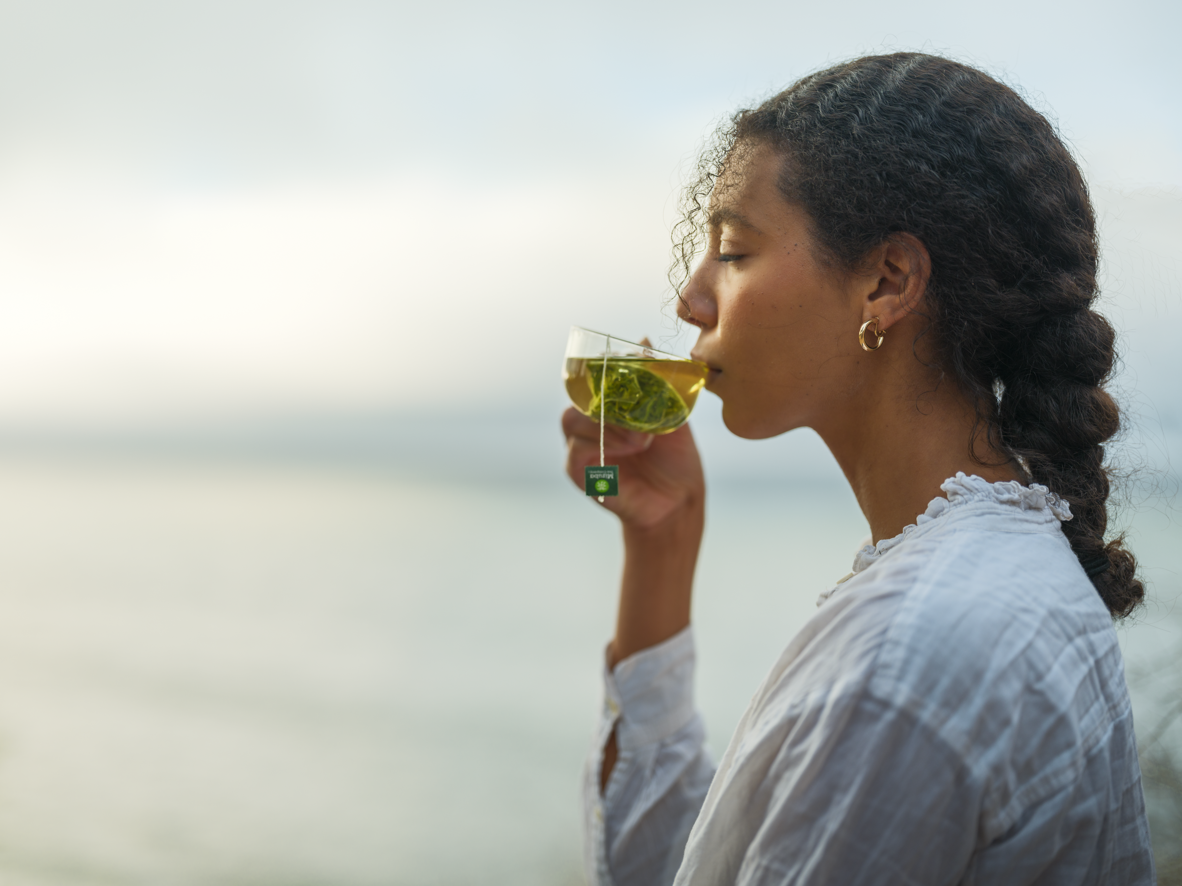 Woman serenely sips from a glass tea cup with a Mizuba Tea Co. organic Kamairicha sachet brewed in it.