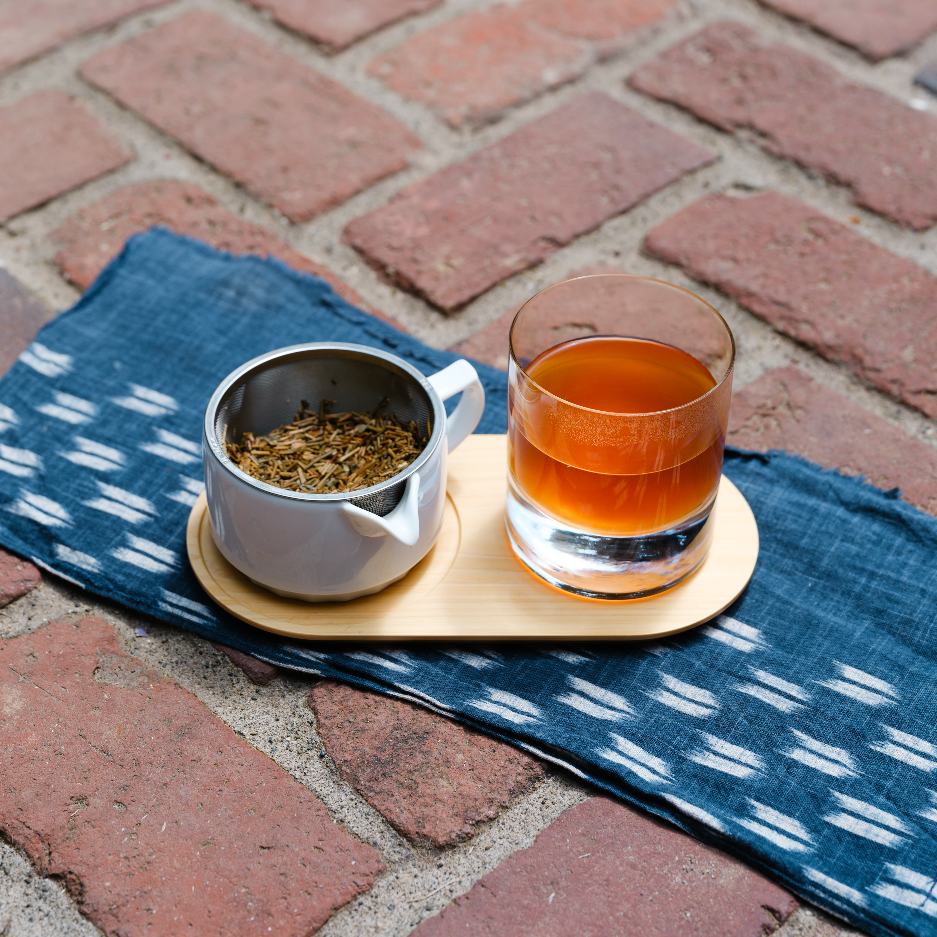 A white teapot steeps Japanese roasted hojicha, which is poured into a glass next to the pot on a tray outdoors
