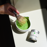Matcha green tea powder being whisked in a chawan tea bowl with a chasen matcha whisk, next to a can of Mizuba matcha powder.