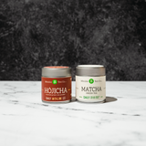 Two Mizuba tea canisters, one labeled 'Hojicha' and the other 'Matcha', on a marble surface with a dark textured background.