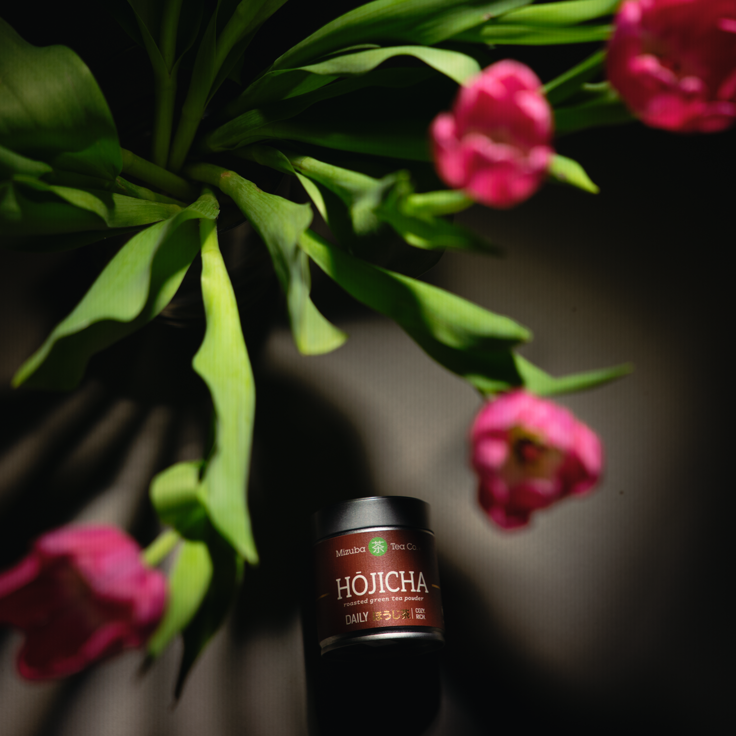Jar of Mizuba Hojicha powder with pink flowers in the background.